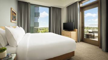A hotel room with a large bed, a flat-screen TV, a small table, and a view of the city through the windows.