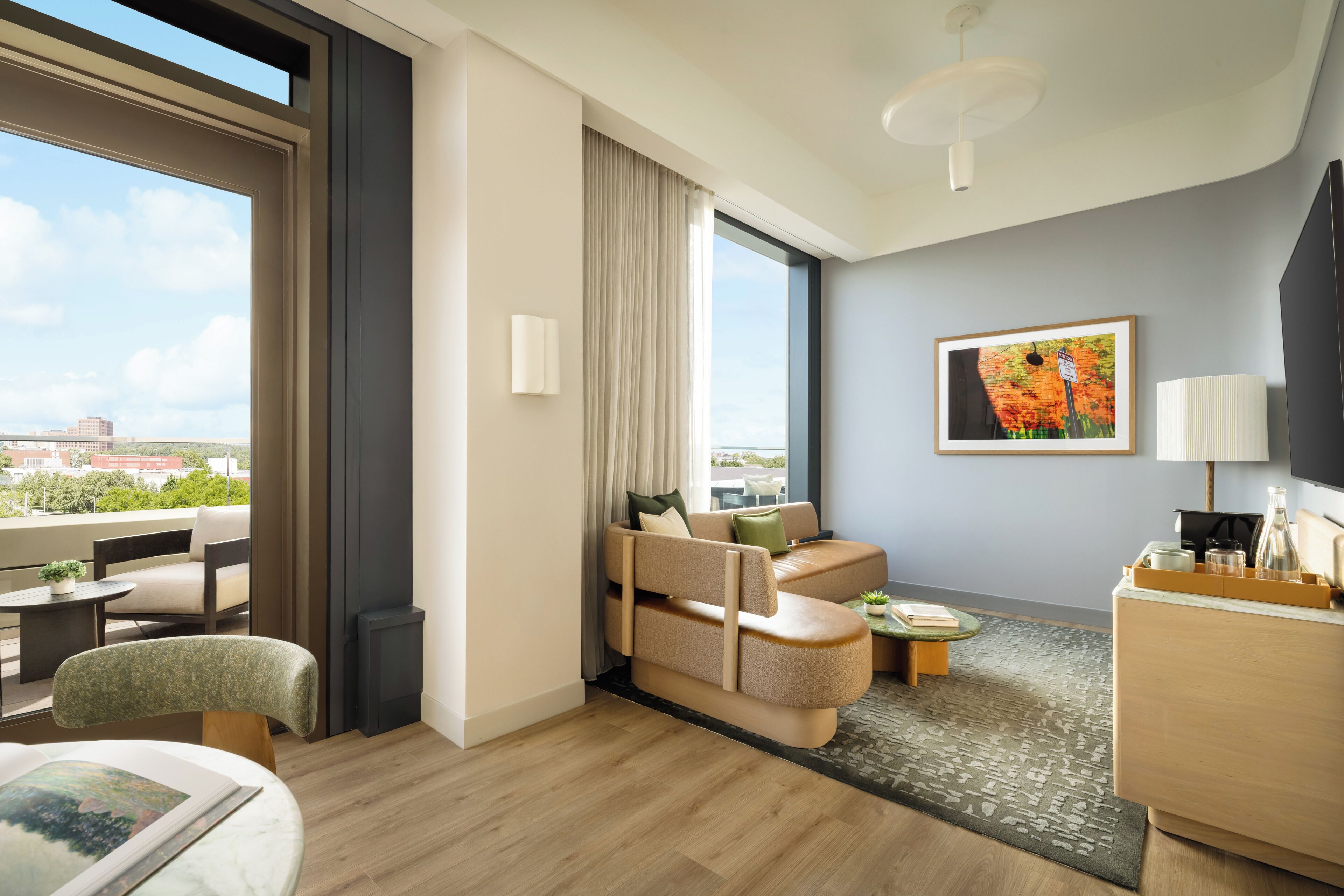 Ein modernes Hotelzimmer mit Sofa, einem kleinen Tisch, einem Schreibtisch, einem Sessel, einem Bett und einem großen Fenster mit Blick auf die Stadt.
