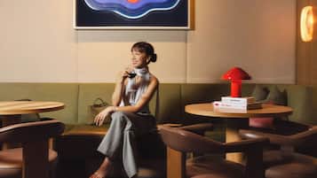 A woman seated on a sofa in a room with a round table, a red lamp, and a Bauhaus-branded box.