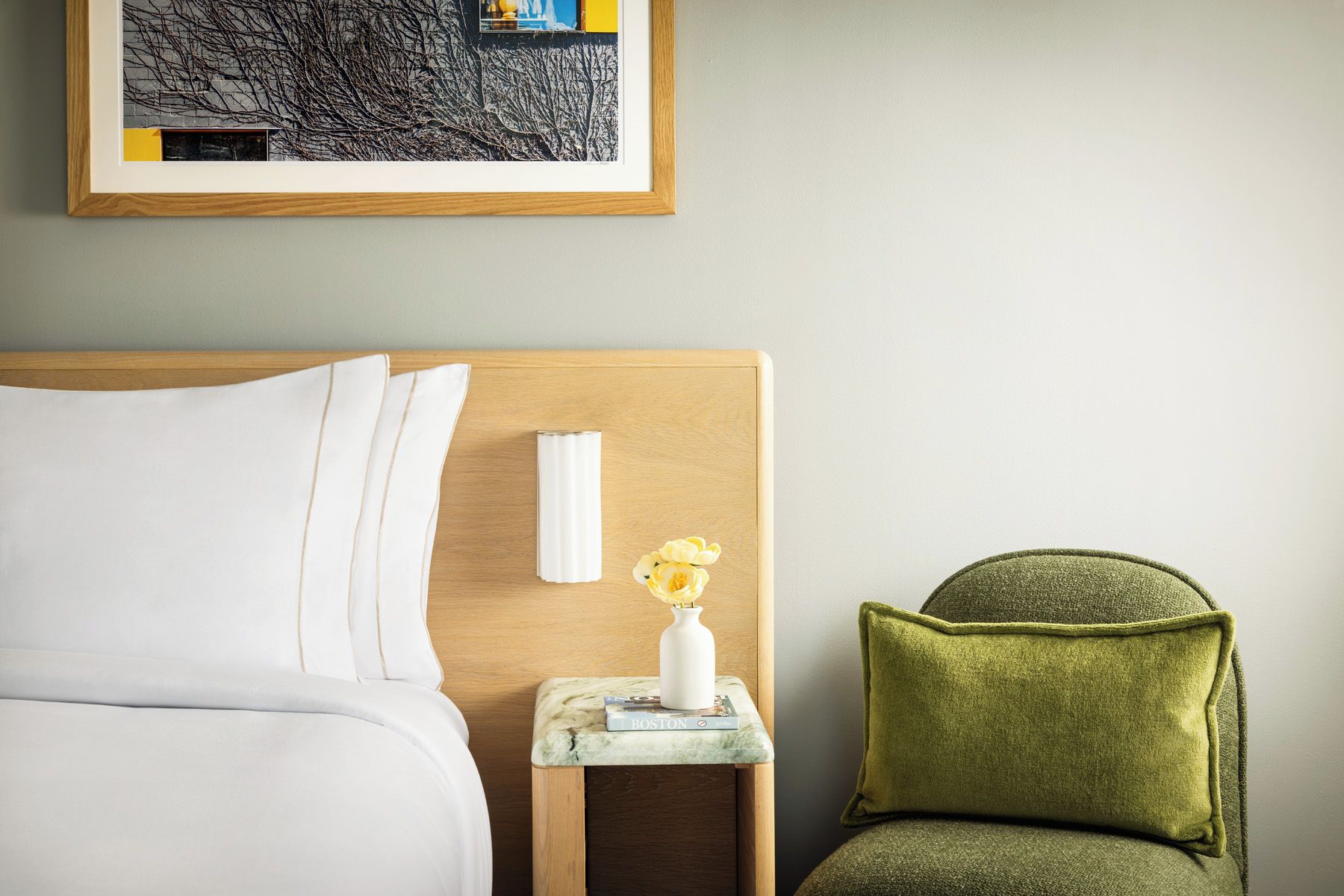 A hotel room with a bed, a nightstand, a vase with flowers, and a green armchair.
