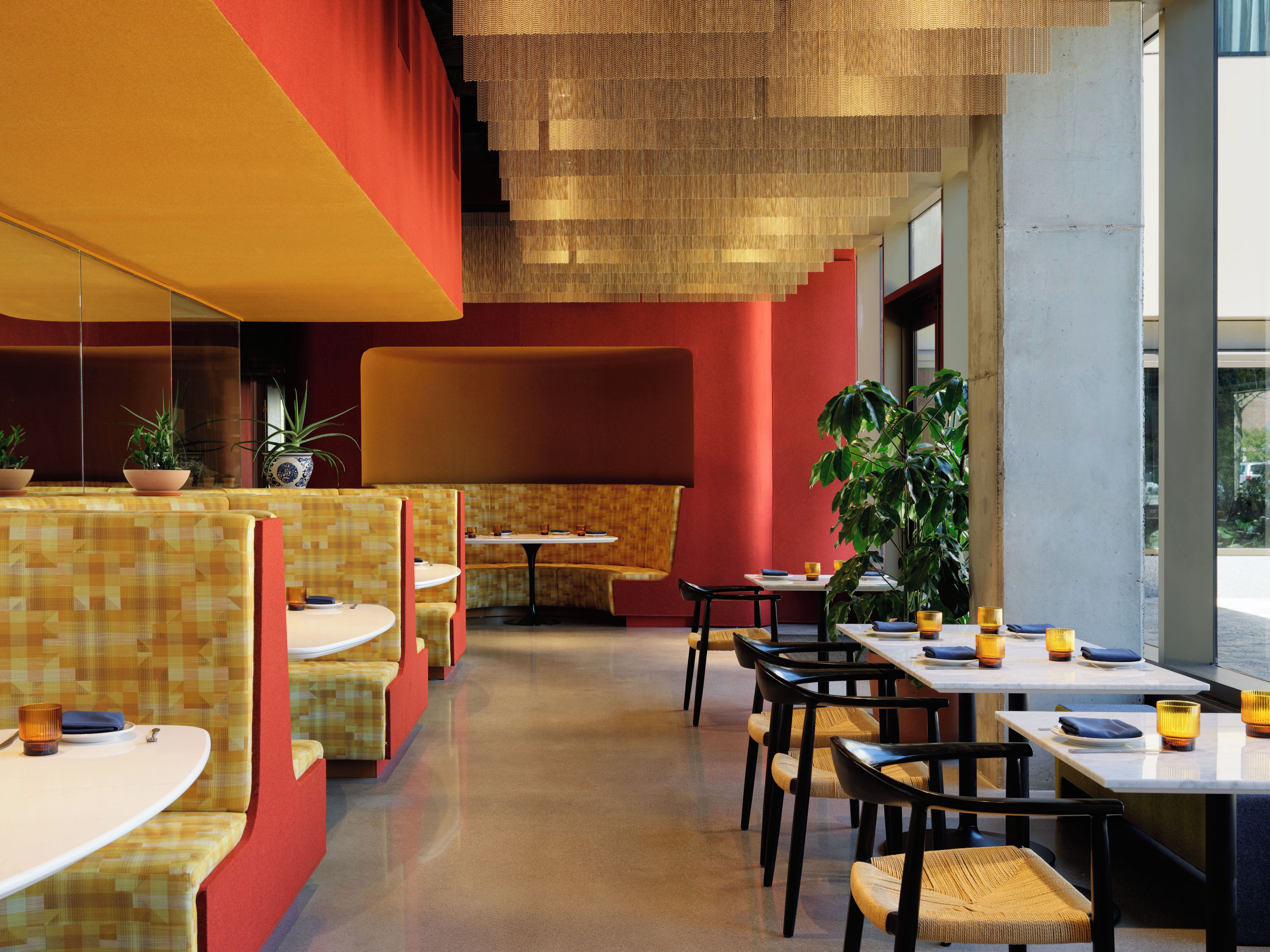 A modern restaurant interior with booths and tables, a large plant, and a red accent wall.