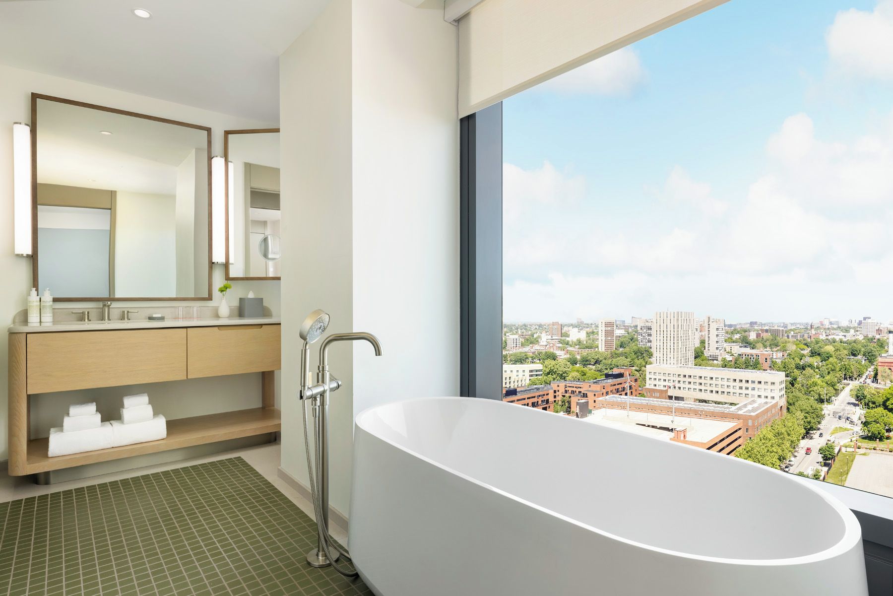 Un baño moderno con una bañera independiente, un espejo grande y vistas a un paisaje urbano a través de un ventanal.