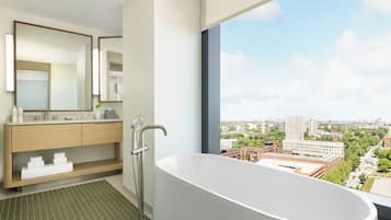 Un baño moderno con una bañera independiente, un espejo grande y vistas a un paisaje urbano a través de un ventanal.