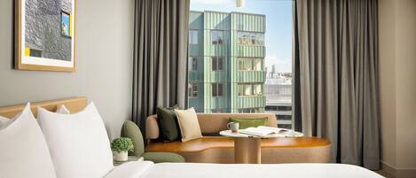 Habitación de hotel con cama, zona de estar con mesa, un cuadro en la pared y vista a un edificio.