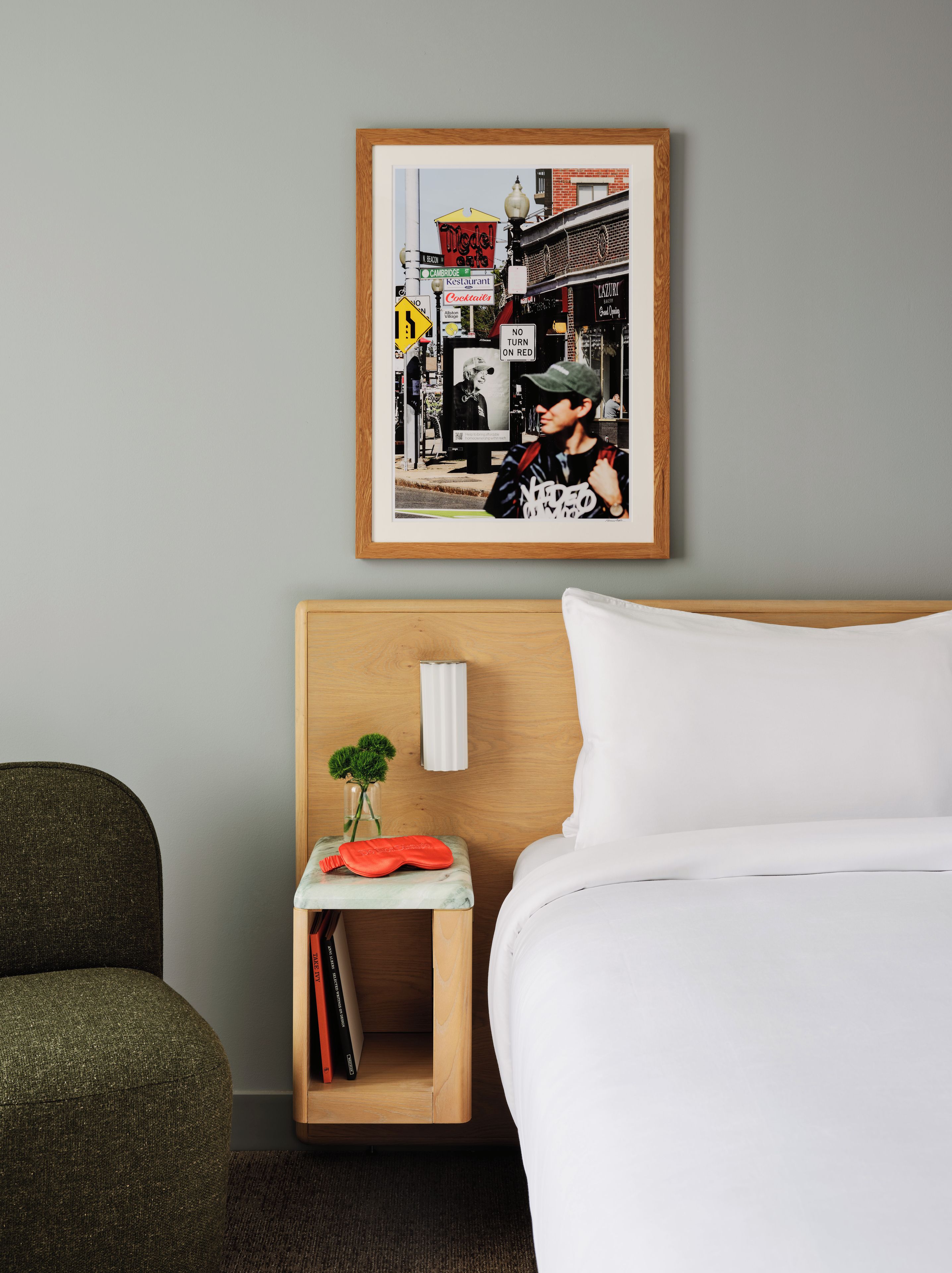 Ein gerahmtes Schwarz-Weiß-Foto einer Straßenszene mit einer Person, das über einem Bett mit weißer Bettwäsche und einem Holz-Kopfteil angebracht ist.