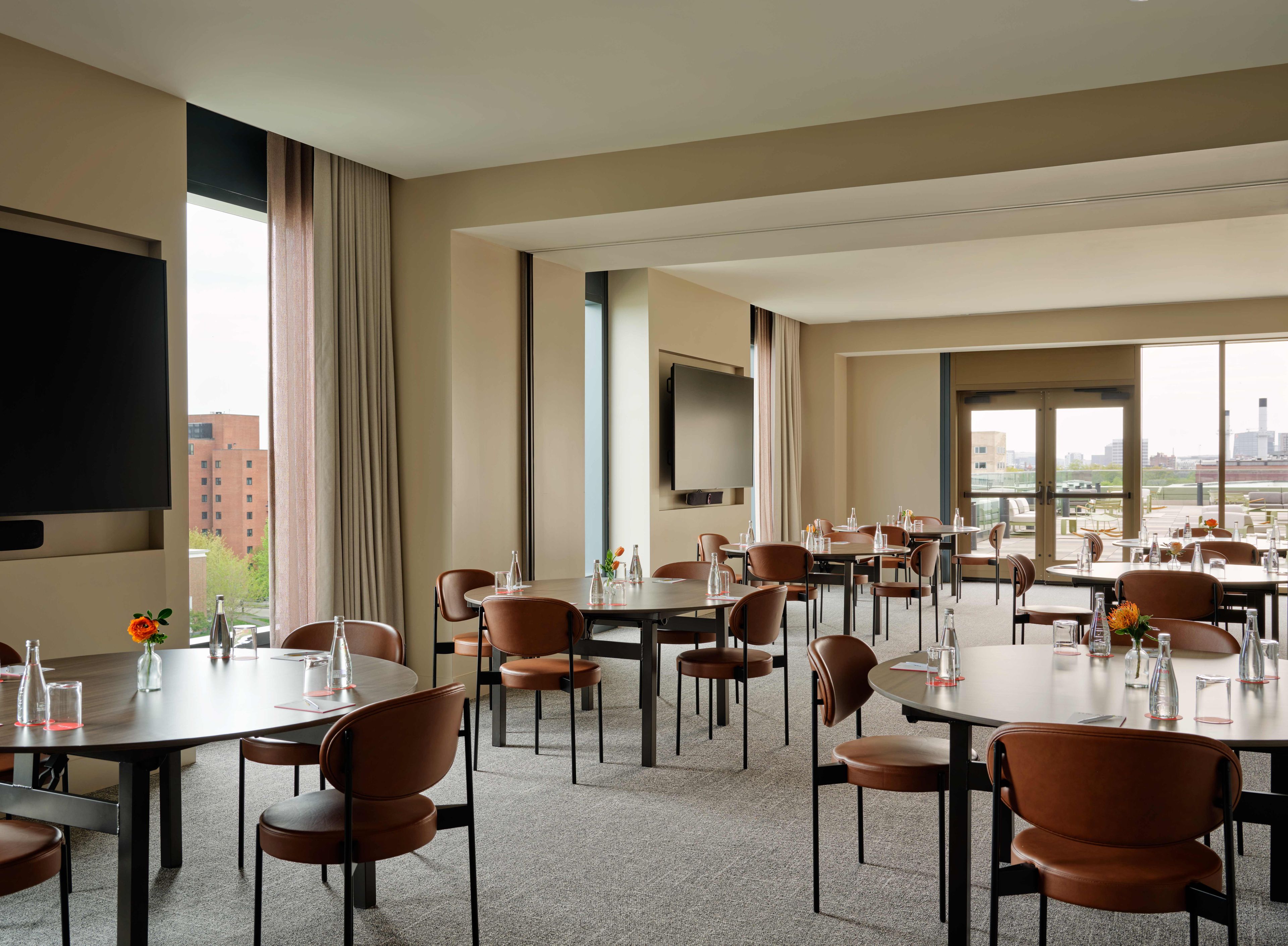 A hotel conference room with multiple tables and chairs, a flat-screen TV, and large windows offering a city view.