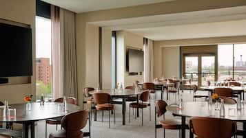 A hotel conference room with multiple tables and chairs, a flat-screen TV, and large windows offering a city view.