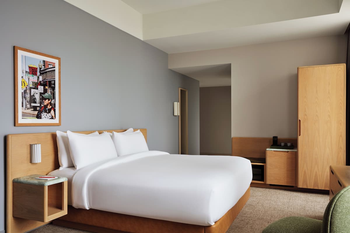Moderni hotellihuone, jossa on suuri sänky, puinen pääty, yöpöytä lampun kanssa, vaatekaappi ja seinällä oleva kehystetty kuva.