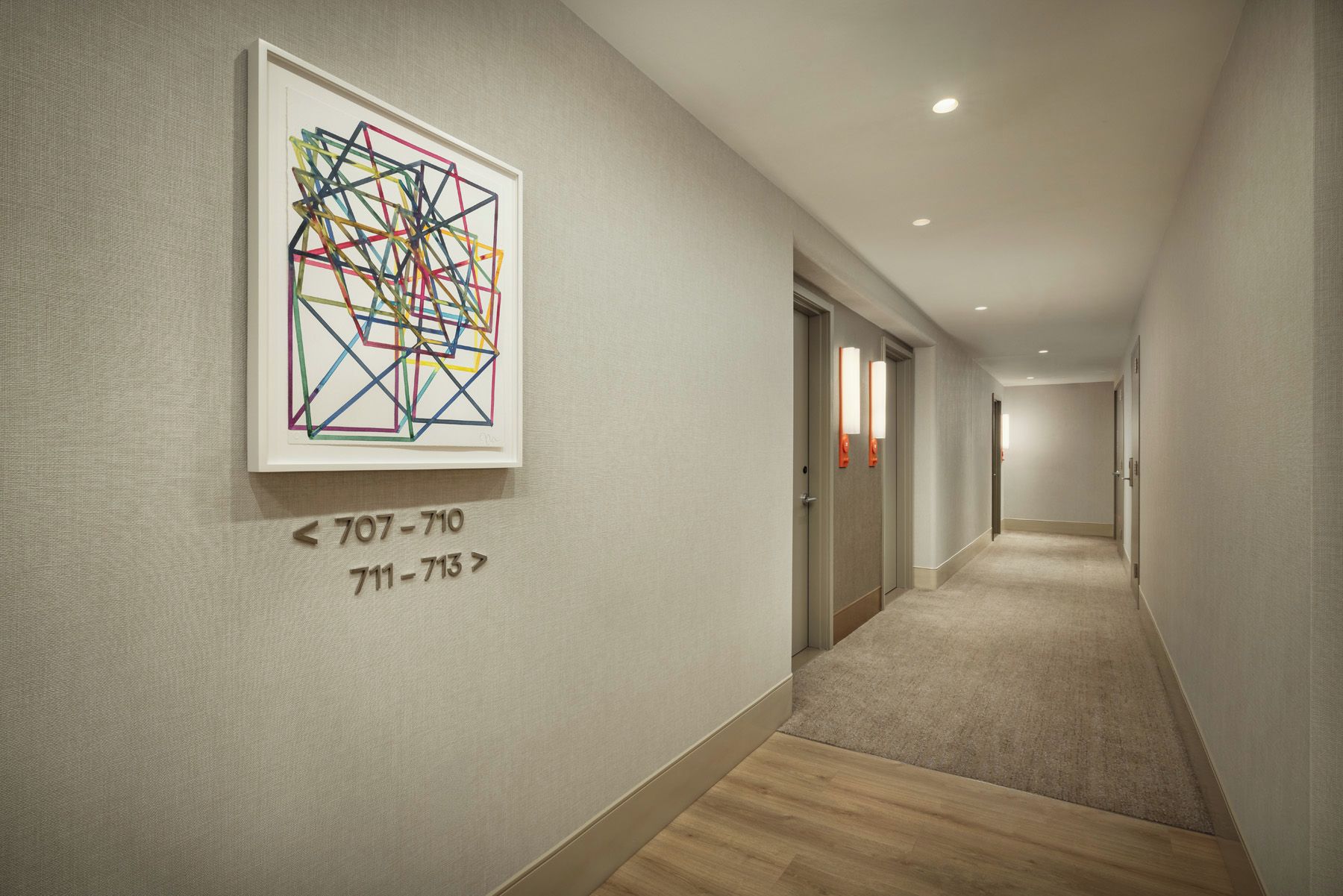 A hotel hallway with a geometric art piece on the wall, carpeted floors, and doors with light fixtures.
