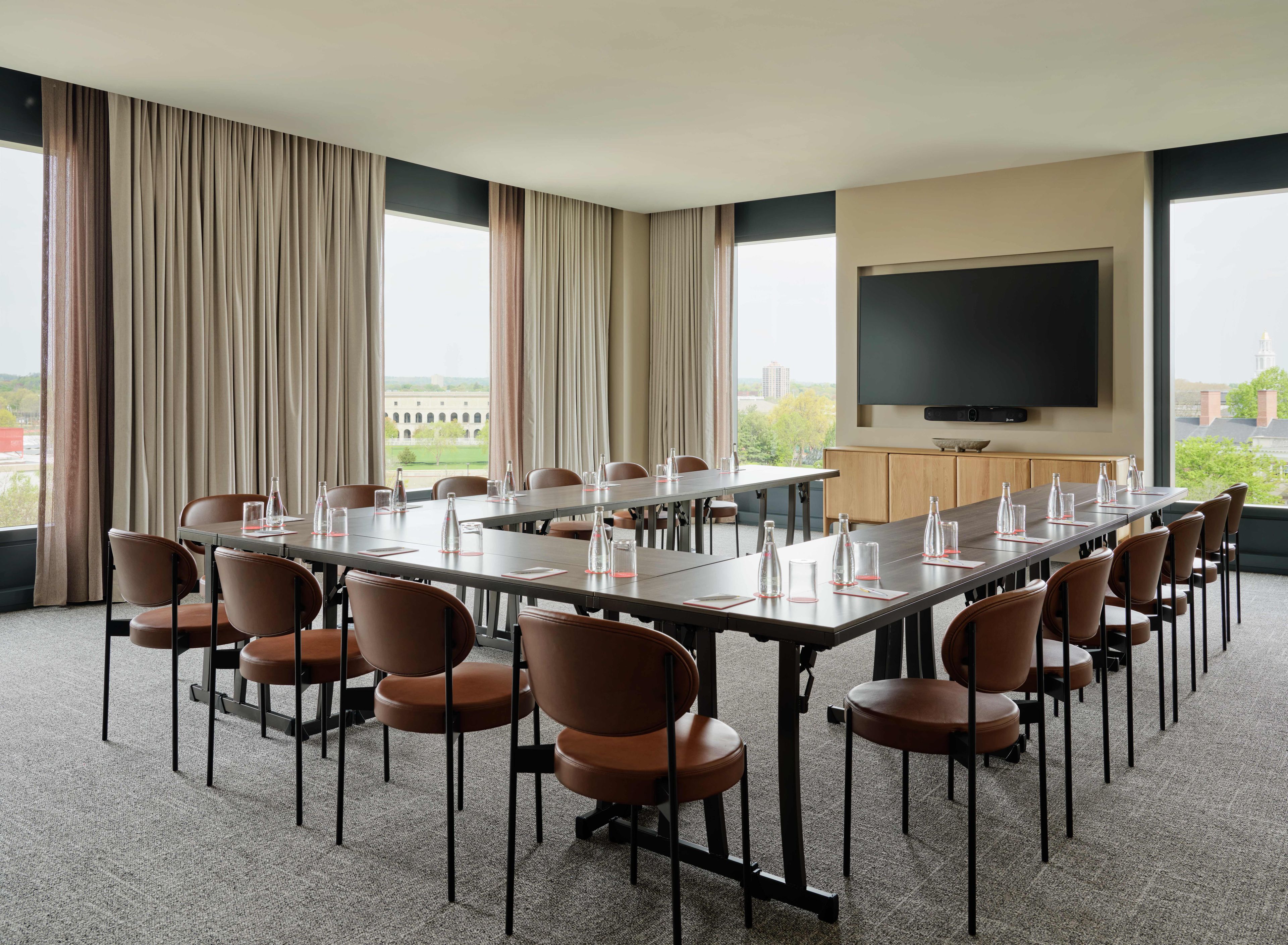 A conference room with a long table, chairs, a flat-screen TV, and large windows with curtains.