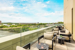 A rooftop terrace with outdoor seating, a glass balustrade, and a view of a building and greenery.