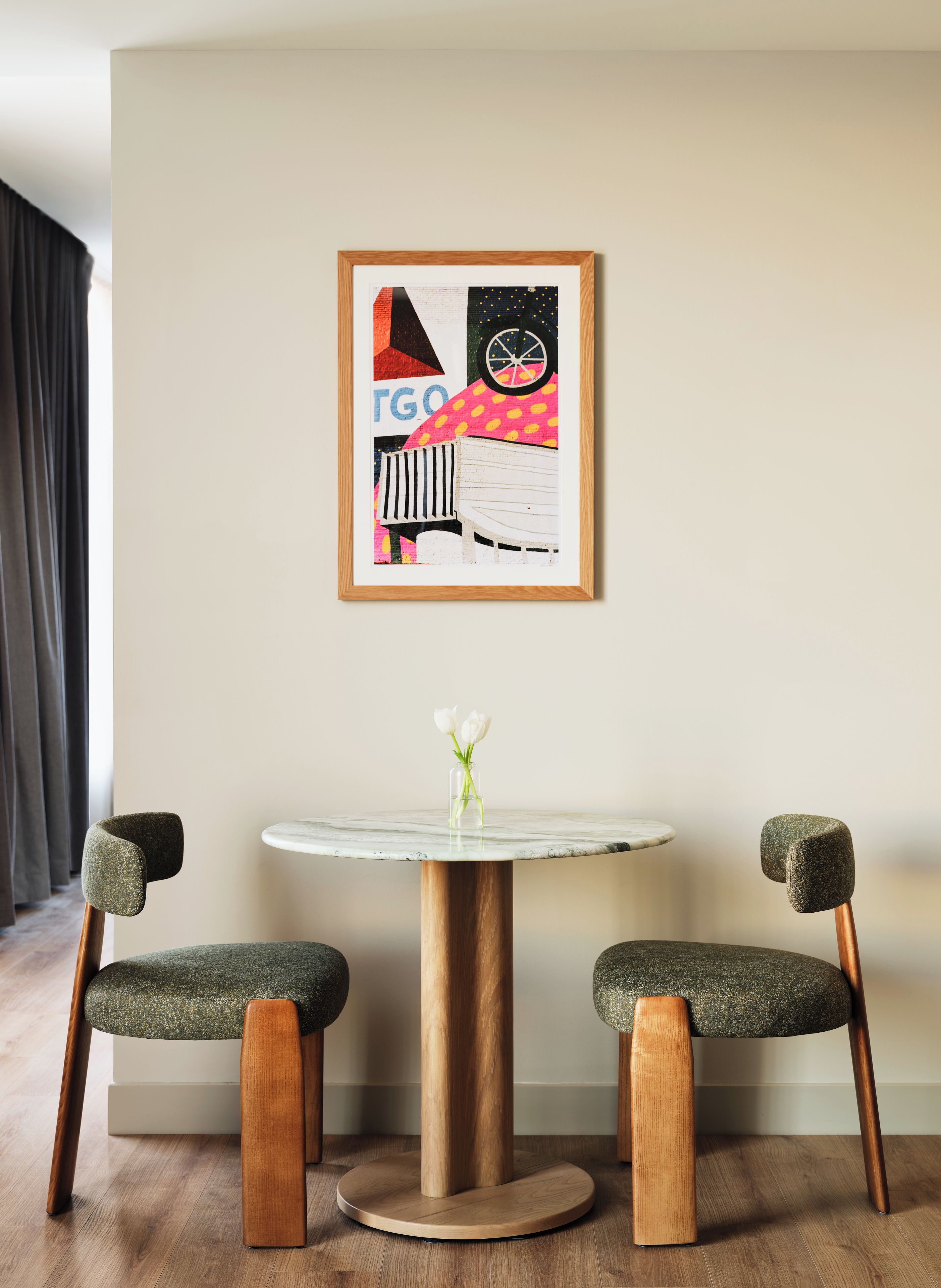 A room with a round table, two chairs, a framed artwork, and a vase with flowers.