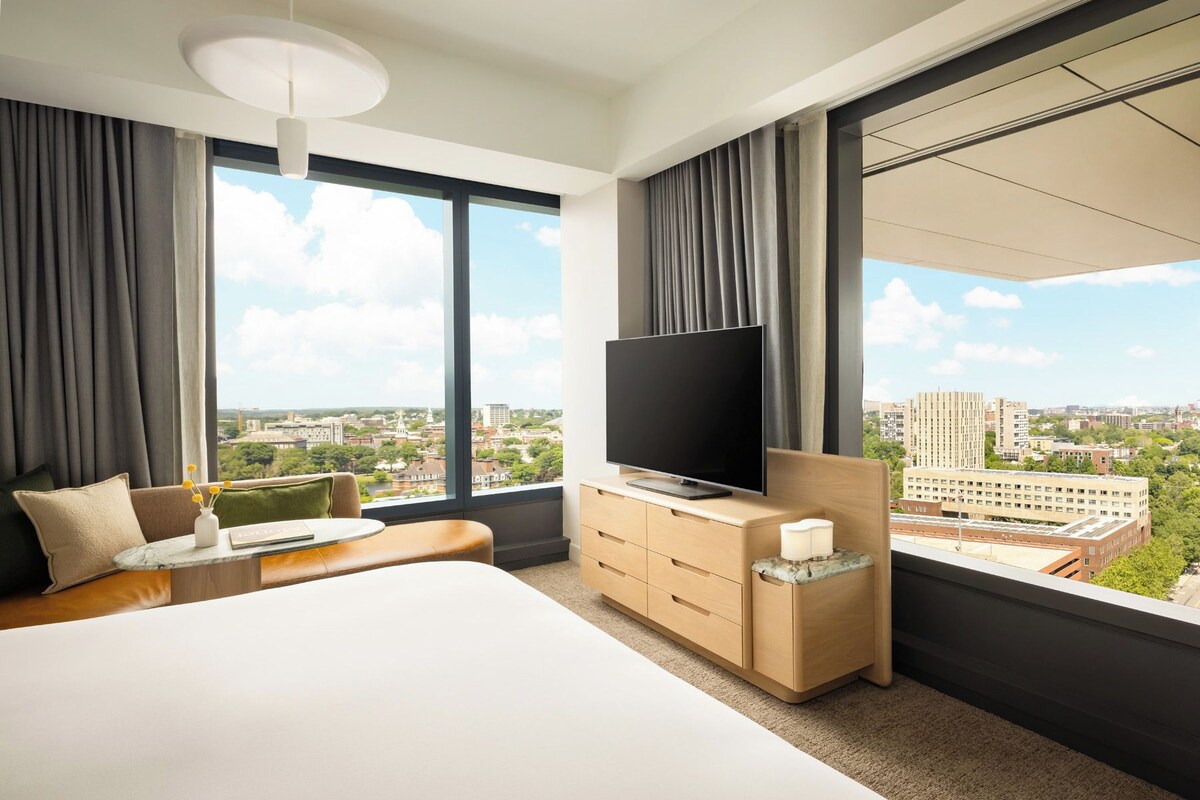 A hotel room with a large bed, a flat-screen TV, a wooden dresser, and a view of the cityscape.