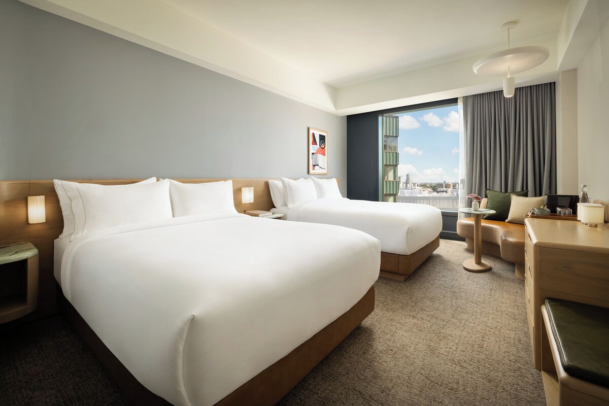 A hotel room with two beds, a desk, a chair, a window with a view, and a lamp.