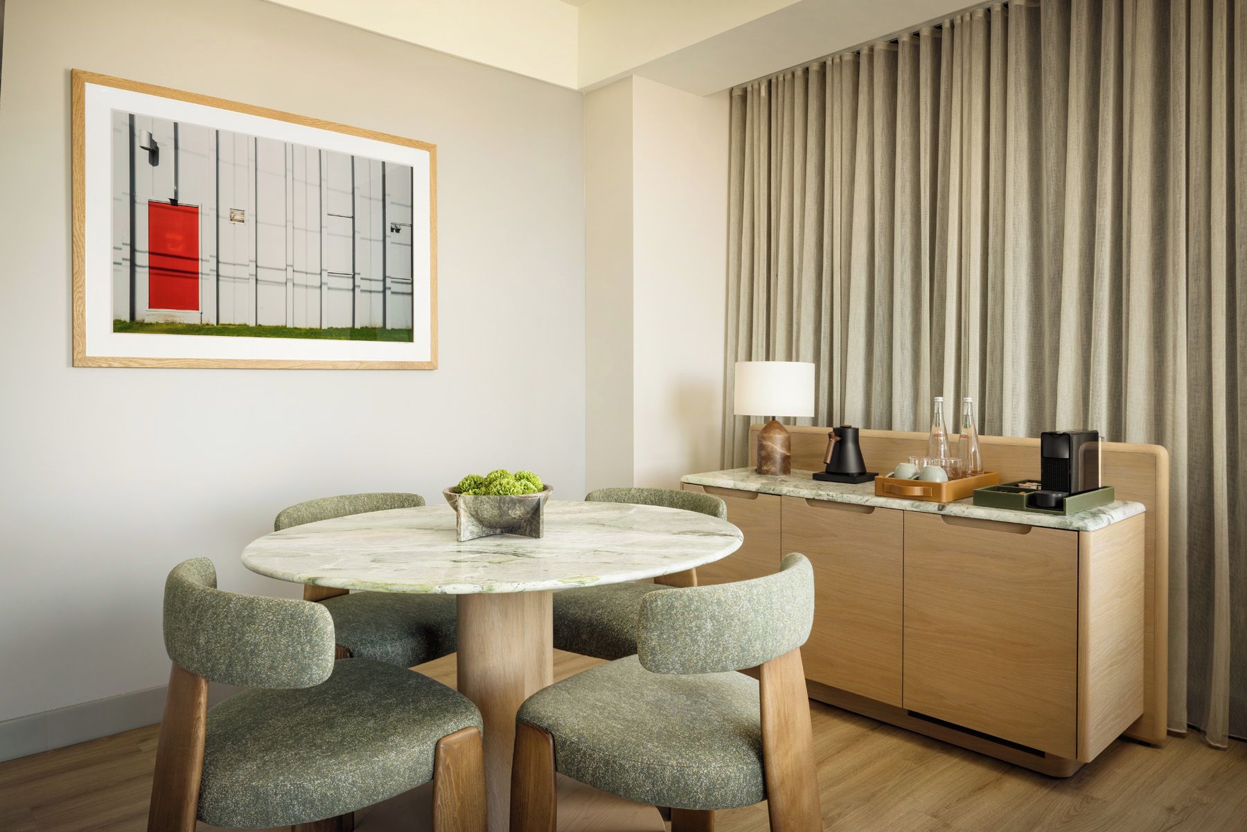A modern hotel room with a round marble table, four upholstered chairs, a wooden cabinet with a countertop, a framed artwork, and a large window with curtains.