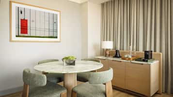 A modern hotel room with a round marble table, four upholstered chairs, a wooden cabinet with a countertop, a framed artwork, and a large window with curtains.