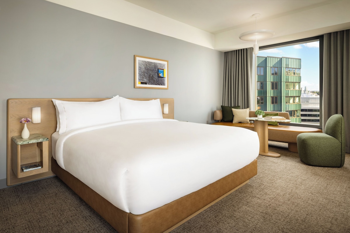 A hotel room with a large bed, a small table, a chair, a nightstand, and a window with a view of a building.