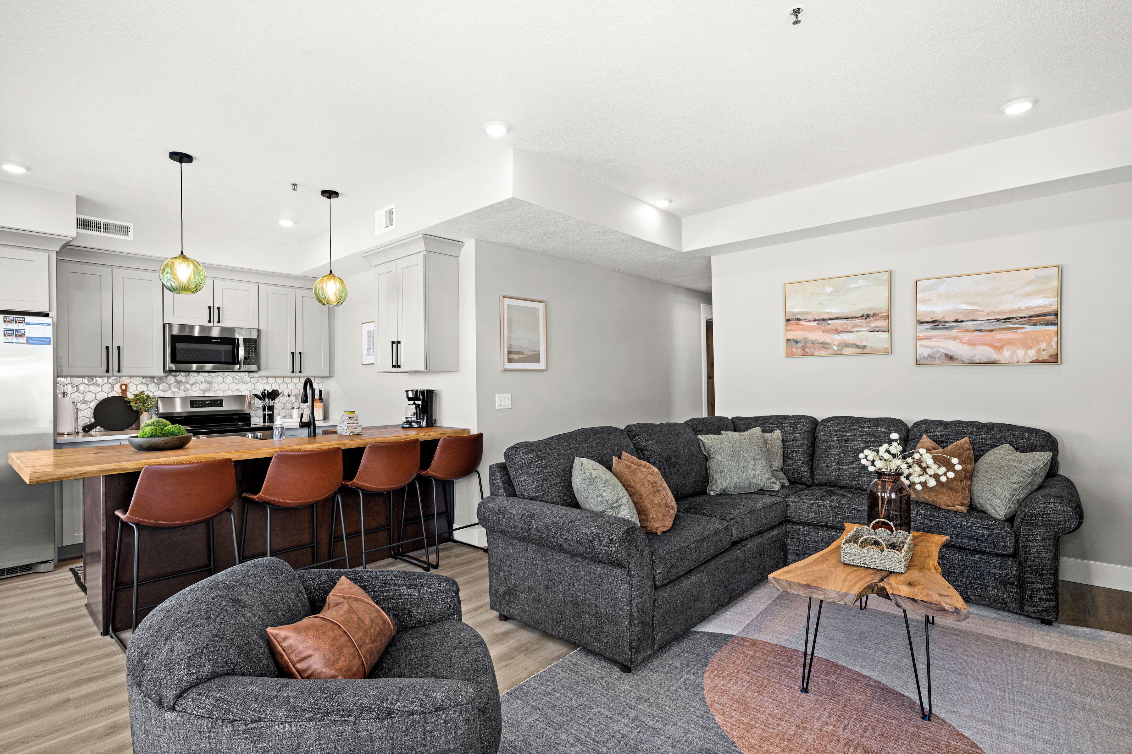 A modern living room with a grey sectional sofa, a wooden coffee table, and a kitchen area with a bar and seating.