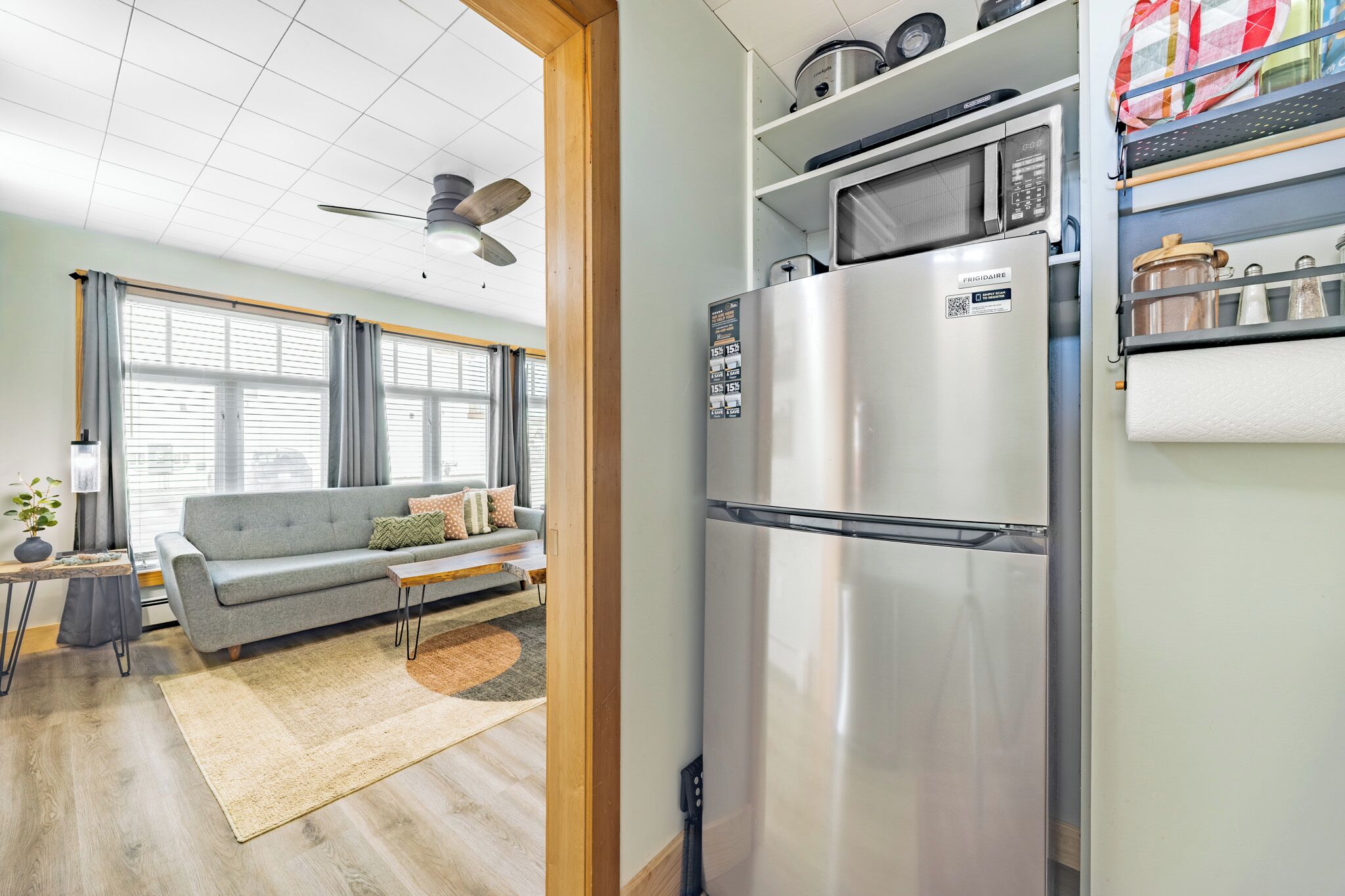 Un espacio habitable pequeño con kitchenette, un sofá gris, un ventilador de techo y una ventana con persianas.