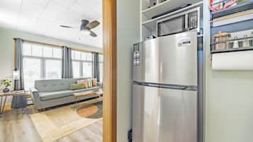 Un espacio habitable pequeño con kitchenette, un sofá gris, un ventilador de techo y una ventana con persianas.