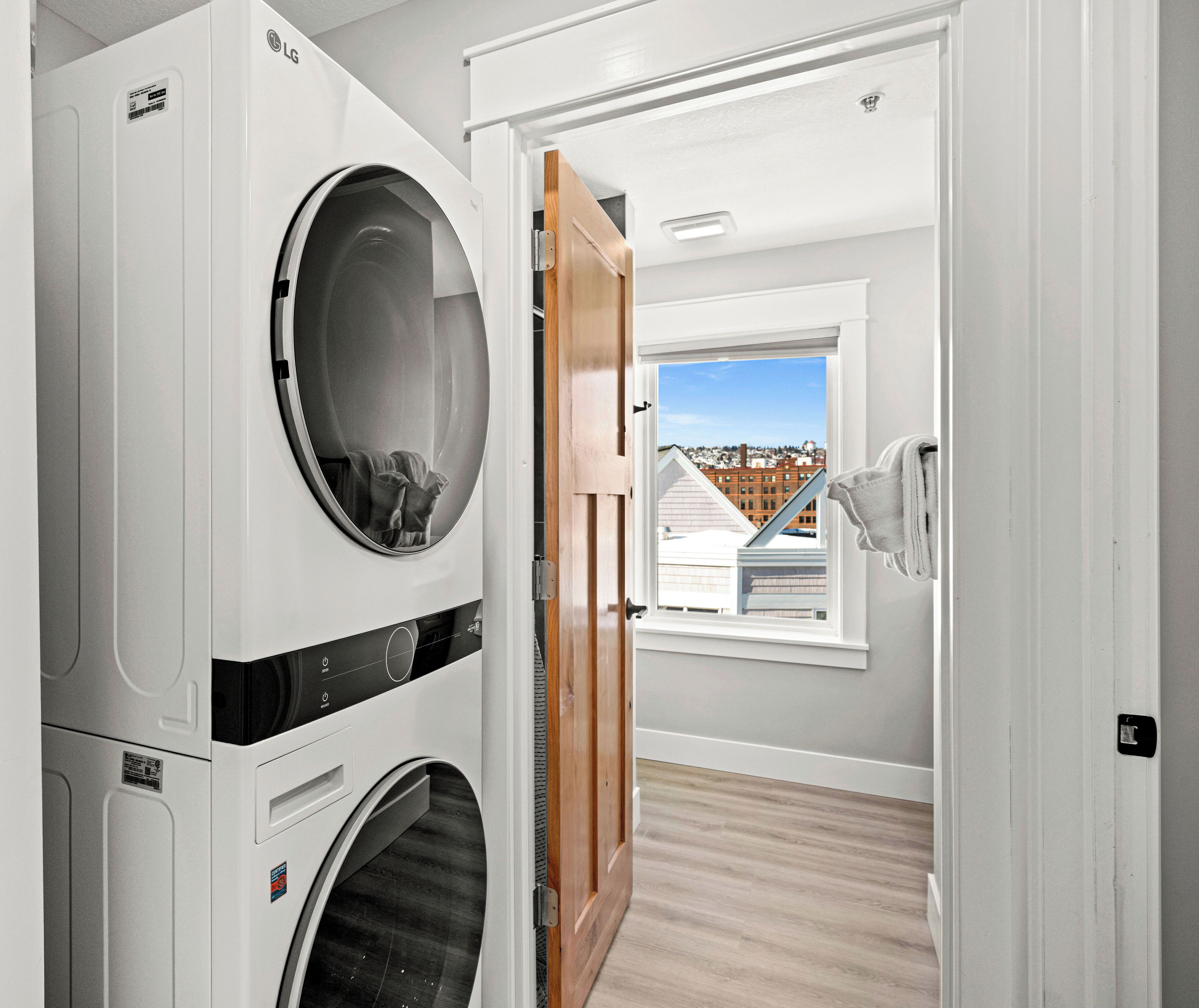 Une buanderie avec des lave-linge et des sèche-linge superposés, une porte en bois donnant sur une pièce avec une fenêtre et une vue sur les bâtiments.