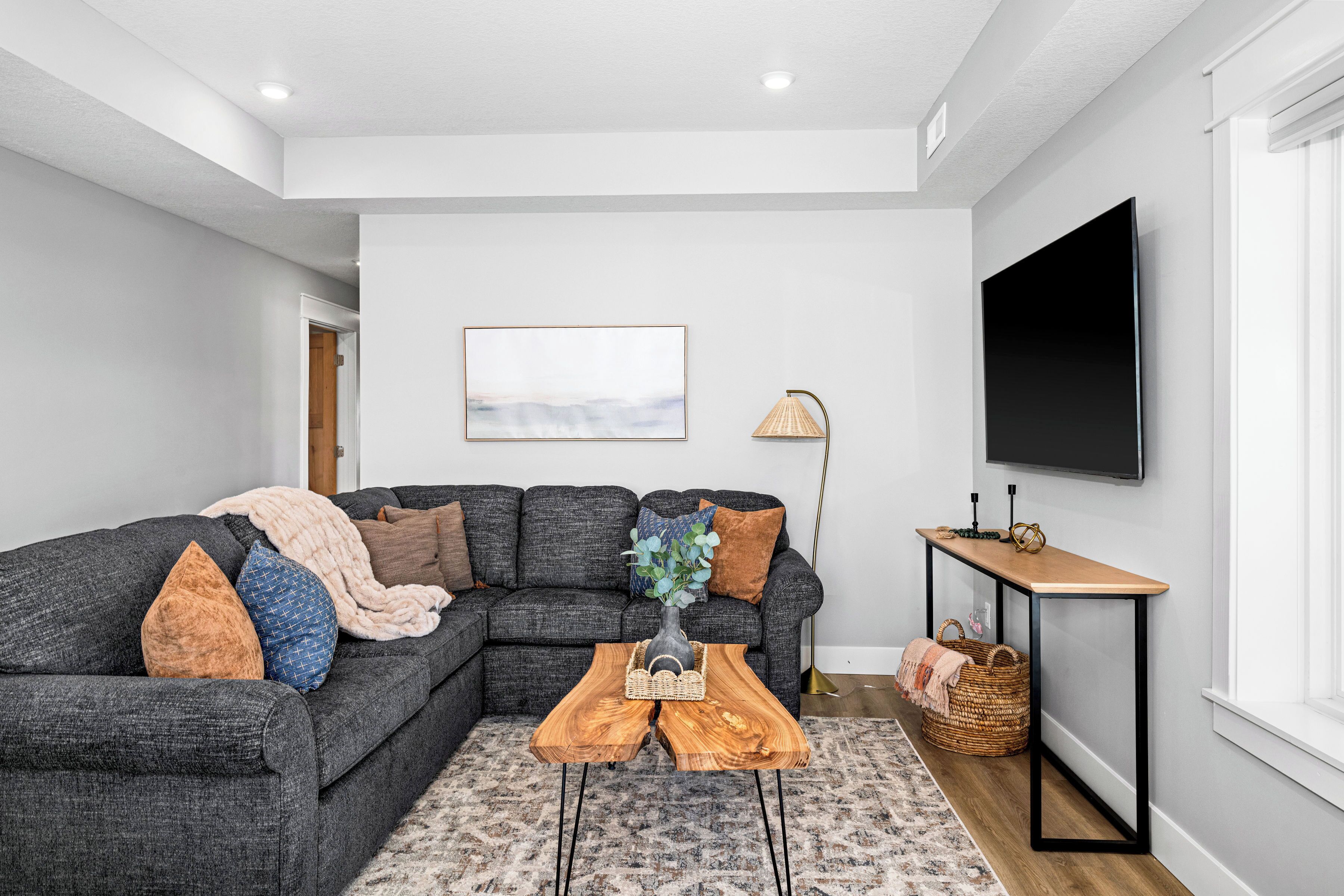 Un salon moderne avec un canapé d’angle gris, une table basse en bois, un téléviseur fixé au mur et une lampe sur pied.