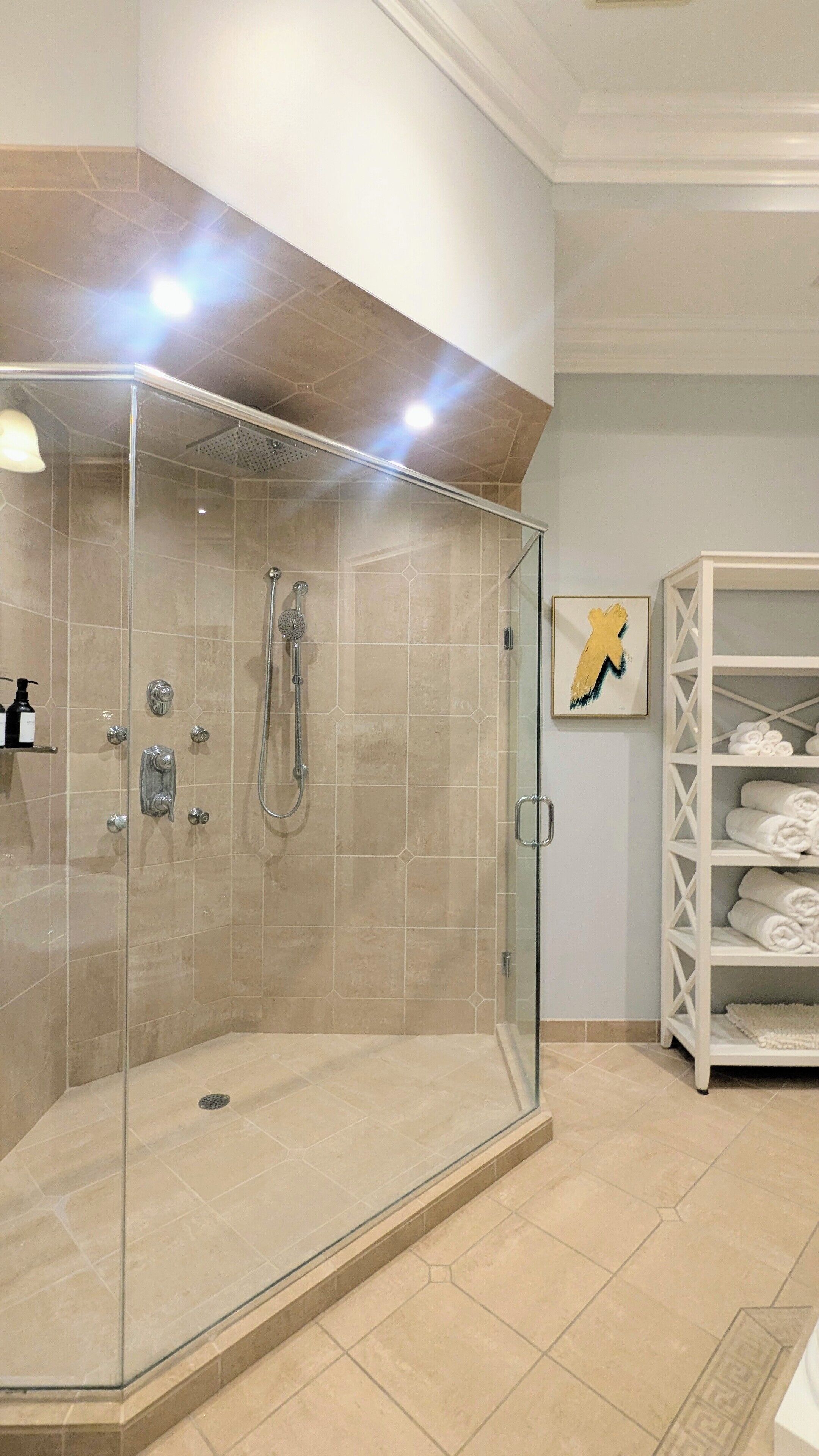 Un baño moderno con ducha acristalada, azulejos color beige, un estante blanco con toallas y una obra de arte enmarcada en la pared.