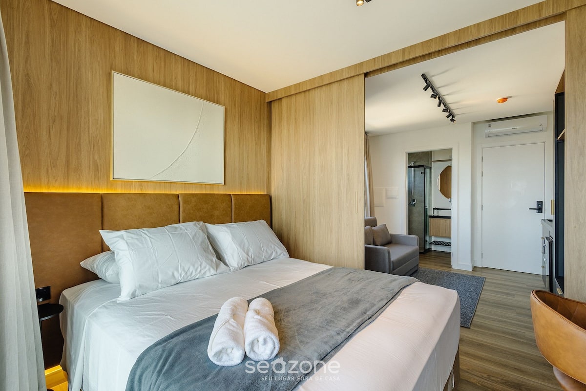 A modern hotel room with a large bed, a headboard, a bedside table, and a view into the adjacent room.