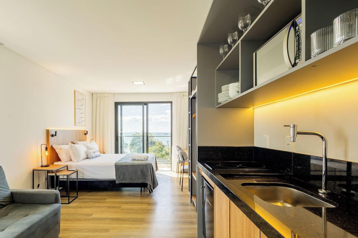 A modern hotel room with a bed, a sofa, a dining table, a kitchen area, and a large window offering a view.