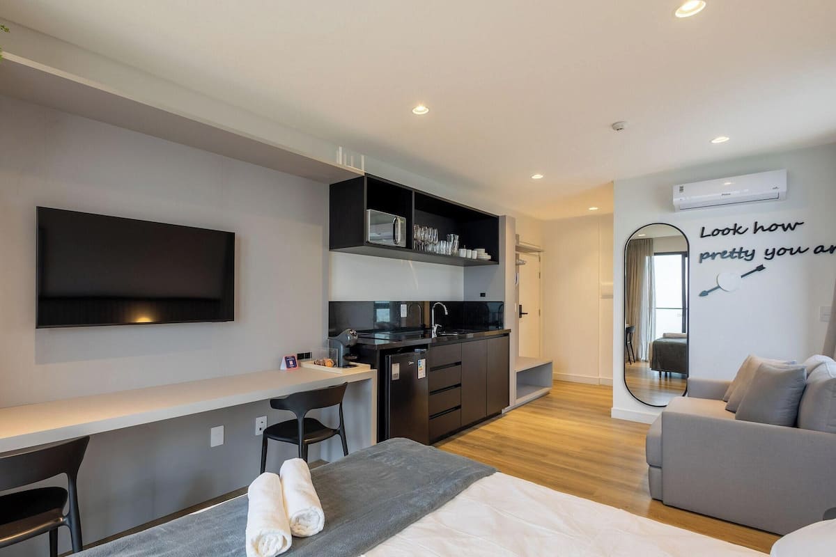 A modern hotel room with a flat-screen TV, a kitchenette, a grey sofa, and a mirror with a motivational quote.
