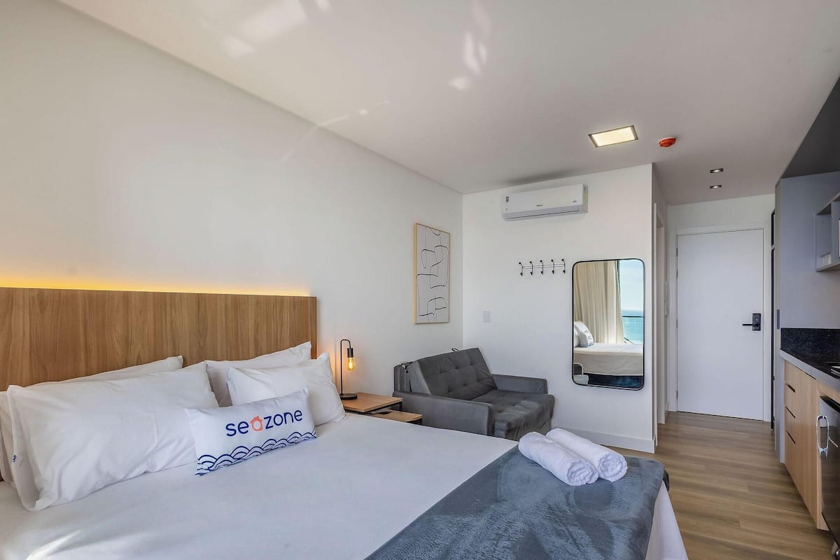 A hotel room with a bed, a desk, a chair, a mirror, and a kitchenette.