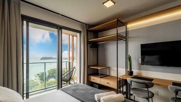 A modern hotel room with a large bed, a desk with a chair, a TV, and a balcony with a view of the sea.