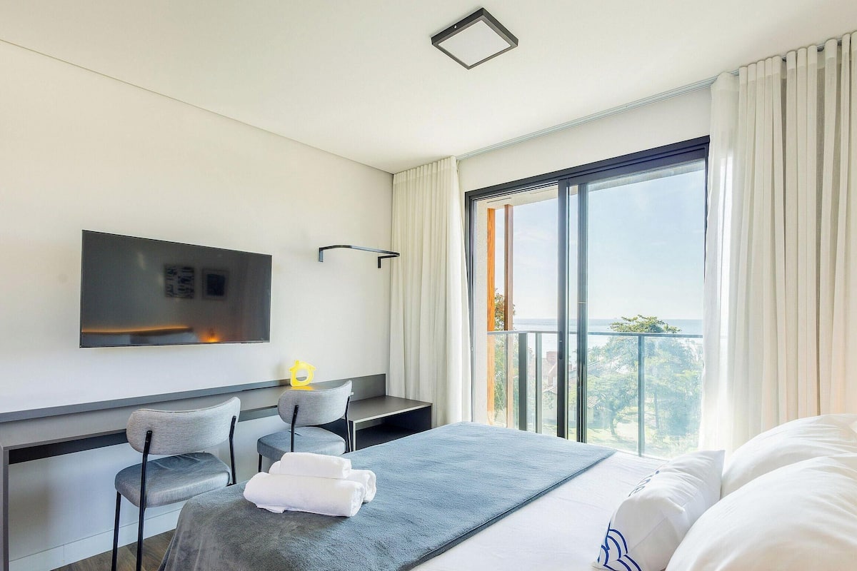 A modern hotel room with a bed, a desk, a chair, a TV mounted on the wall, and a large window with a view.