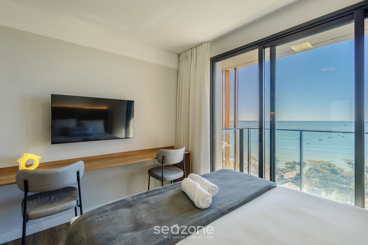 A hotel room with a bed, a TV, a chair, and a large window with a view of the sea.