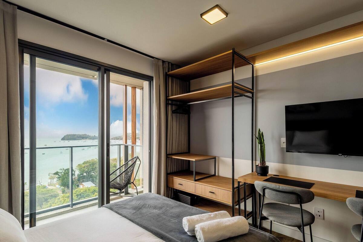 A modern hotel room with a large balcony, a bed, a desk with chairs, and a TV.