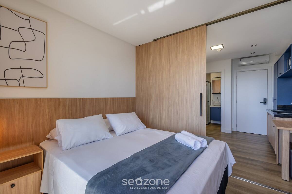 A modern hotel room with a wooden headboard, a large bed with white linens, a grey throw blanket, and a wall-mounted abstract art piece.