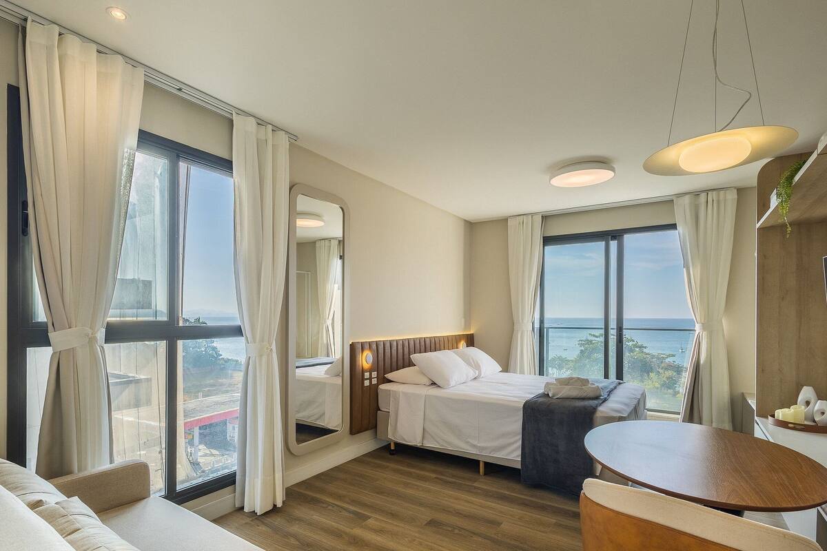 A hotel room with a large bed, a sofa, a dining table, and a view of the sea.