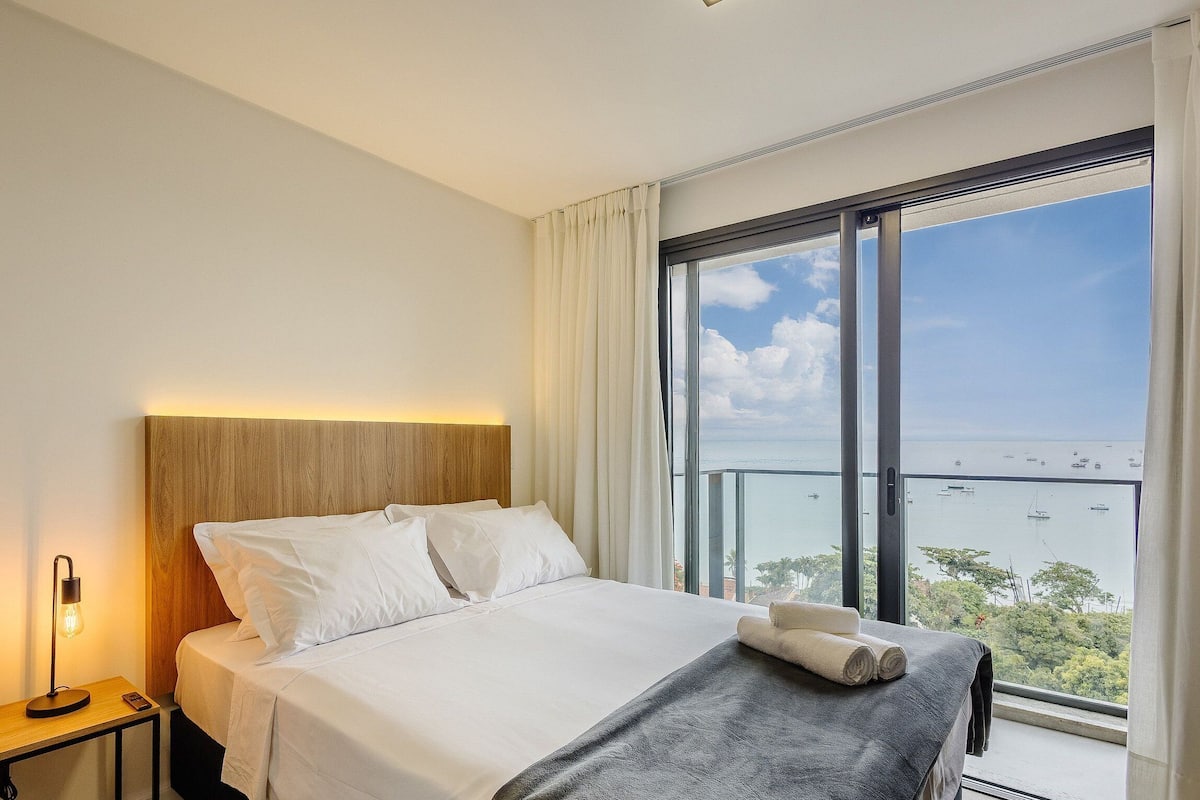 A hotel room with a bed, a nightstand, a lamp, and a large window with a view of the sea.