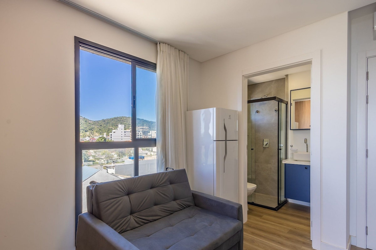 Un apartamento moderno con sofá, refrigerador, ducha de vidrio y vistas a edificios y colinas.