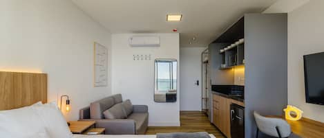 Habitación de hotel compacta con cama, sofá, escritorio pequeño, silla, kitchenette y un ventanal grande.