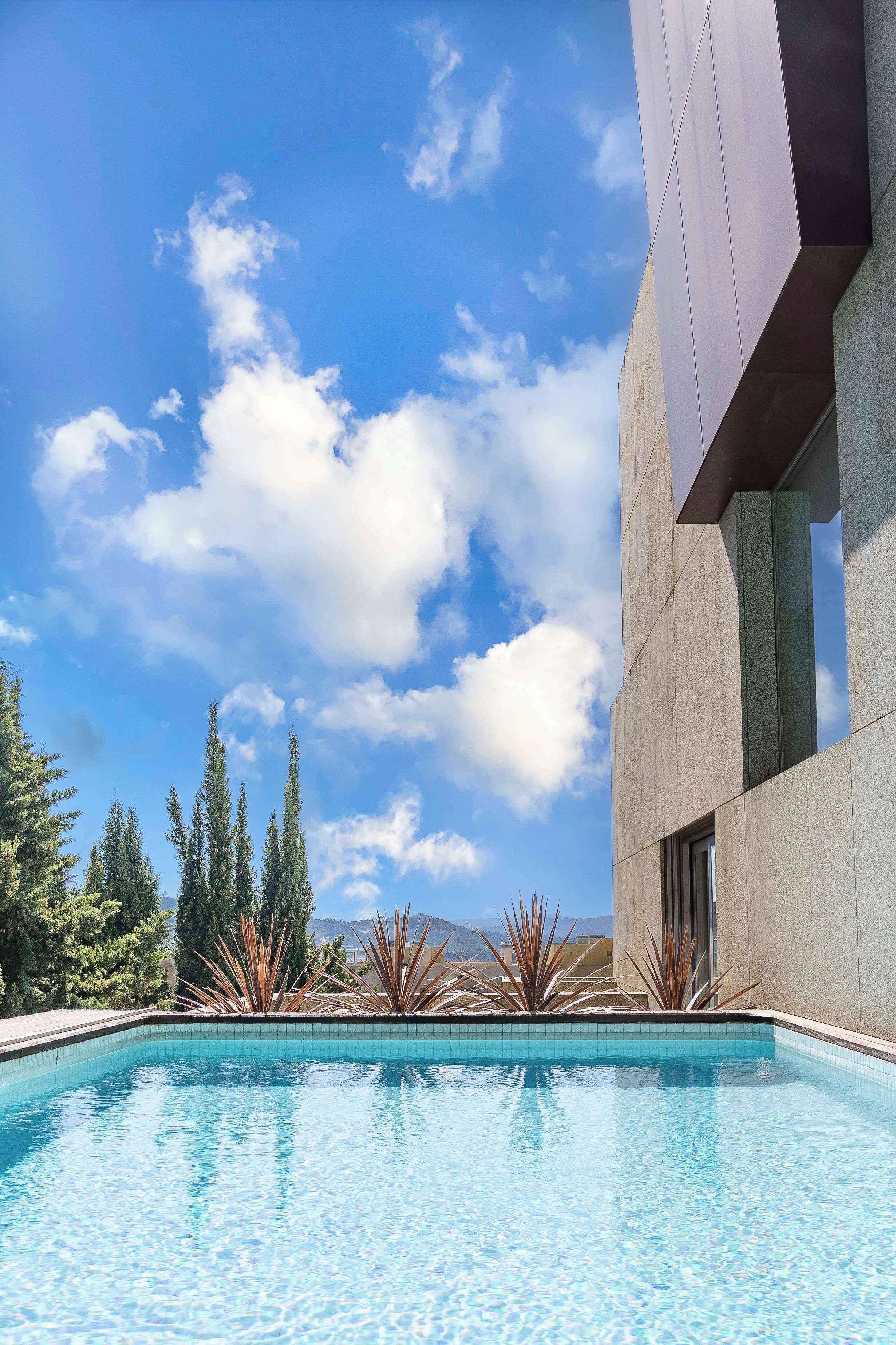 A swimming pool with clear blue water, surrounded by a modern building with a tiled border, and a clear sky with clouds.