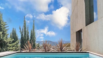 A swimming pool with clear blue water, surrounded by a modern building with a tiled border, and a clear sky with clouds.