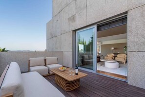 Un área de estar moderna al aire libre con una mesa de madera, sofás acolchados y un ventanal de vidrio que da a un espacio interior con una mesa redonda y sillas.
