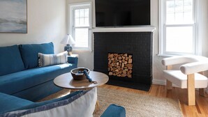 Un salón con un sofá azul, un sillón blanco, una mesa de centro redonda, una chimenea con revestimiento de ladrillo negro y un televisor de pantalla plana montado sobre la chimenea.