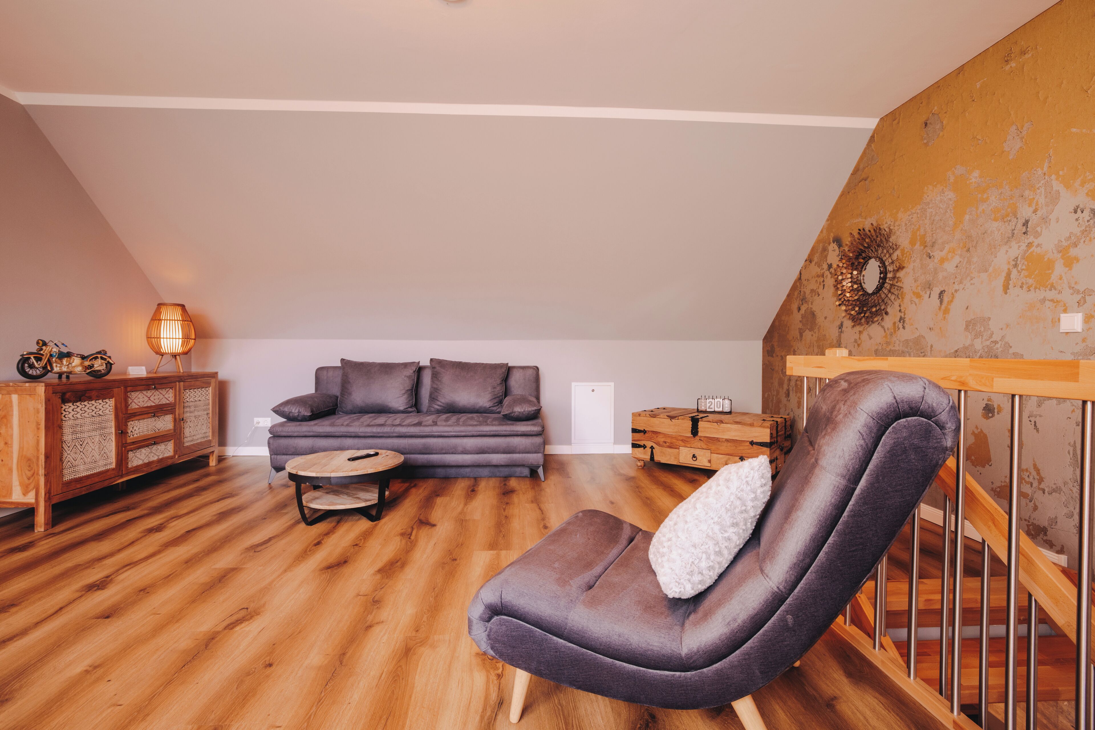 A cozy living room with a wooden floor, a gray sofa, a cushioned armchair, and a small round table.