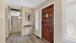 A hallway with wooden flooring, a door with a grid pattern, and a small table with a lamp. - Balloon Fiesta foothills Hidden Gem (Albuquerque)
