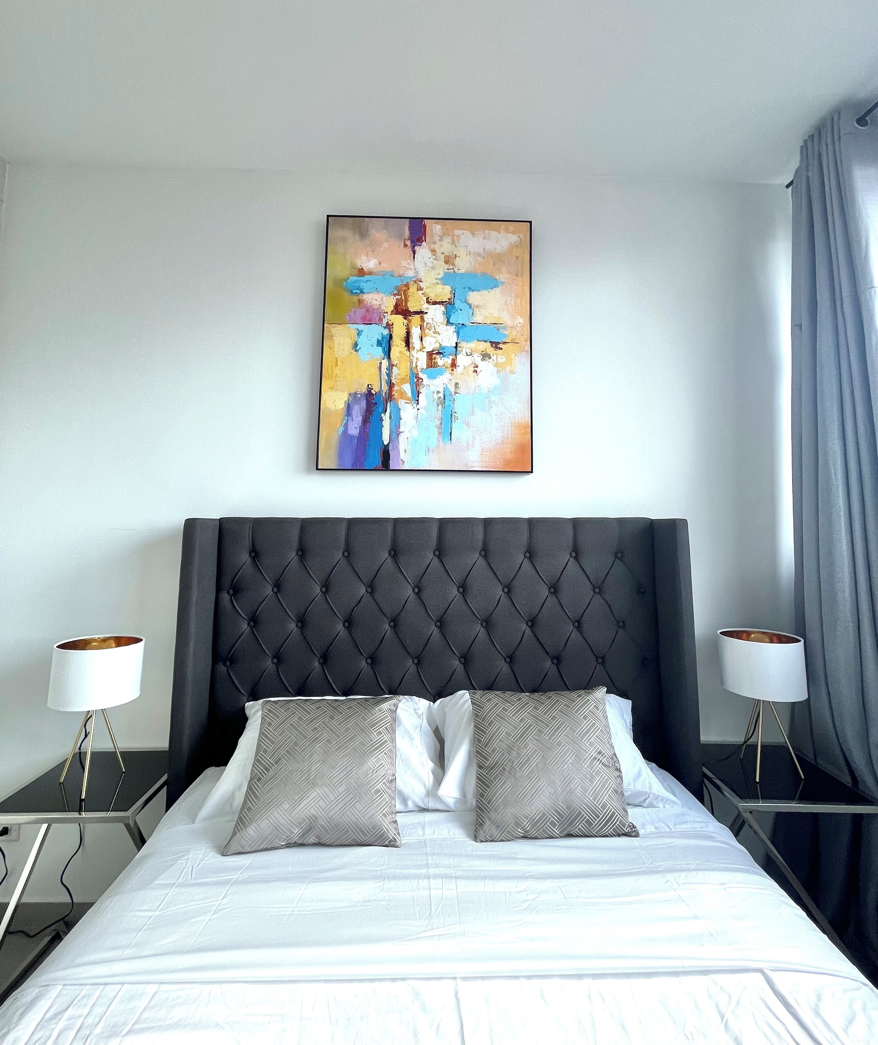 Une chambre d’hôtel moderne avec une tête de lit capitonnée, un grand tableau abstrait, deux tables de chevet avec des lampes et un lit soigneusement fait avec des oreillers à motifs.