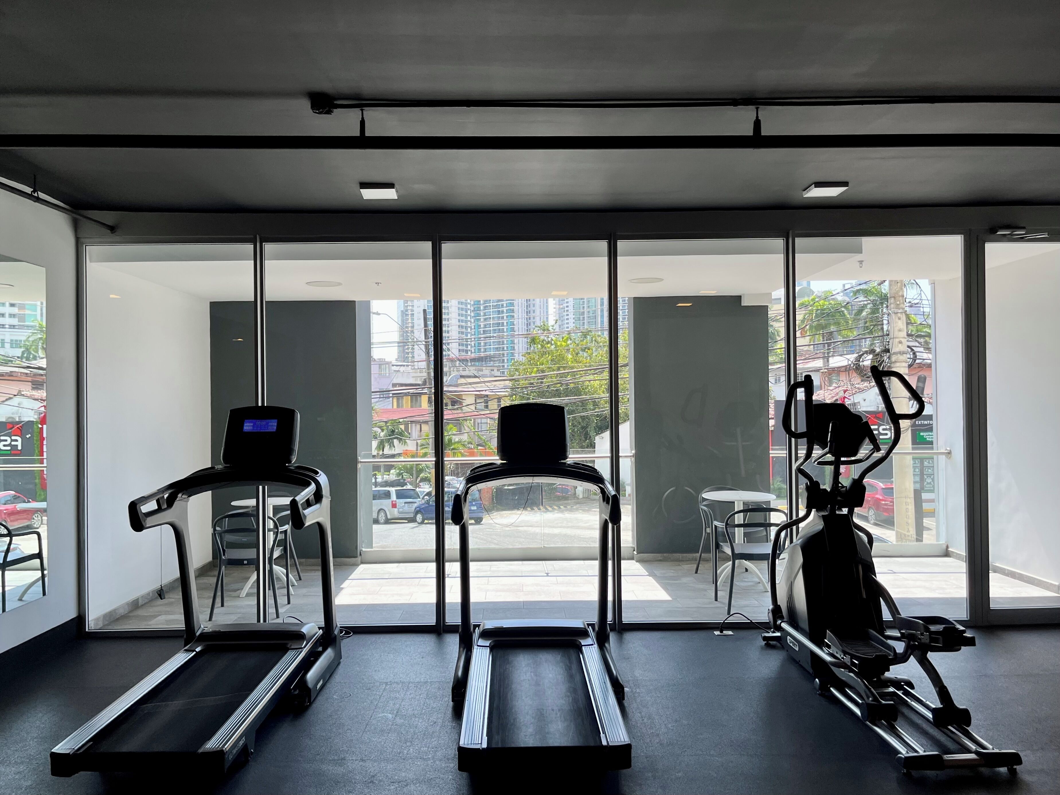 A gym with treadmills and elliptical machines, large windows with a city view, and a balcony with outdoor seating.