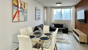 A modern living room with a glass dining table, white chairs, a black sofa, and a TV mounted on the wall.
