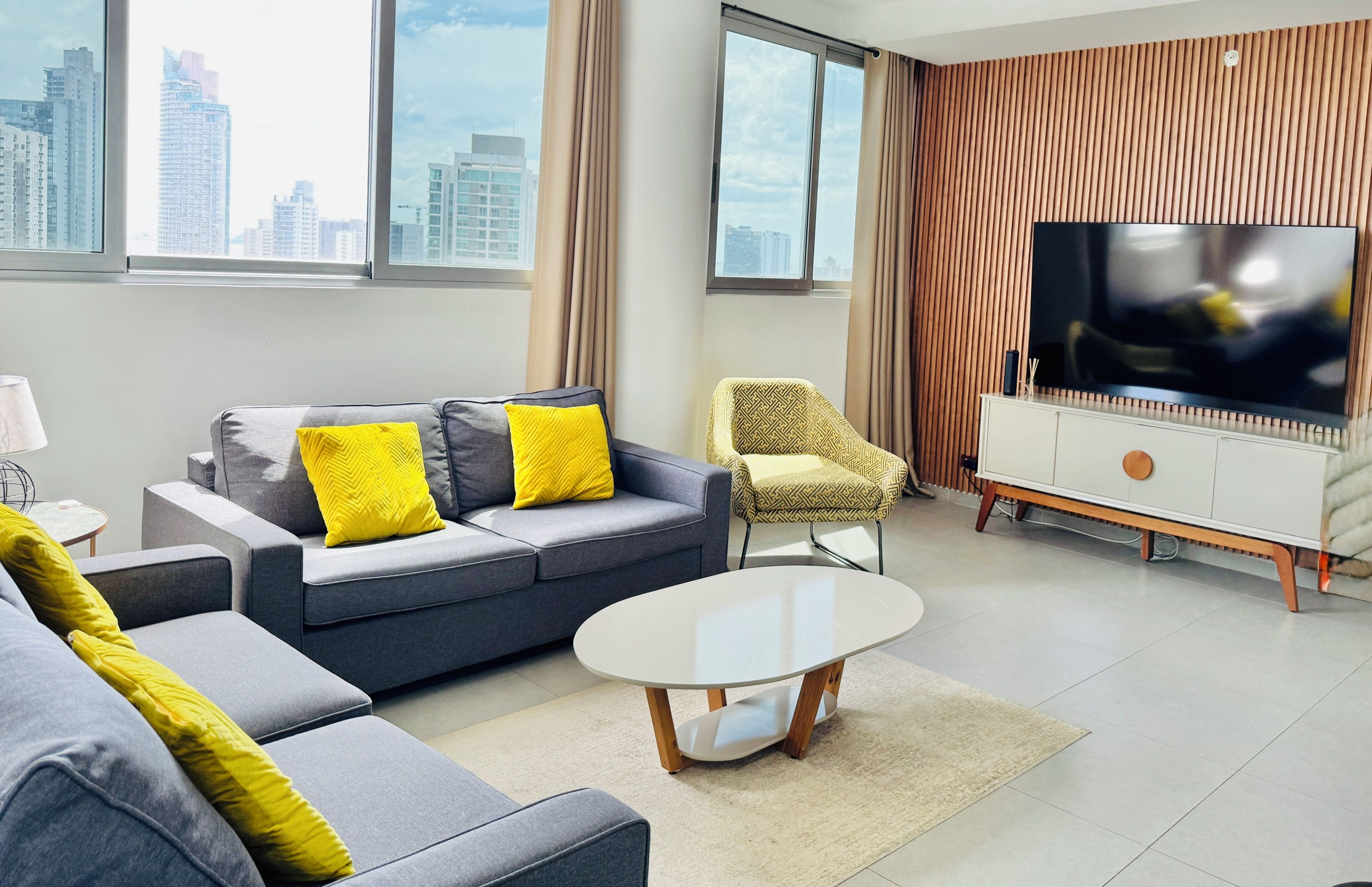 Una sala de estar moderna con un sofá gris, cojines amarillos, una mesa de centro redonda, un sillón estampado y un televisor grande sobre un mueble de madera.